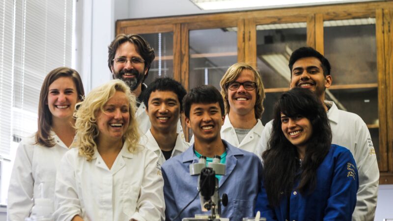 A photograph of a group of sudents in lab coats behind a microscope in a lab, with their bearded advisor standing behind them.