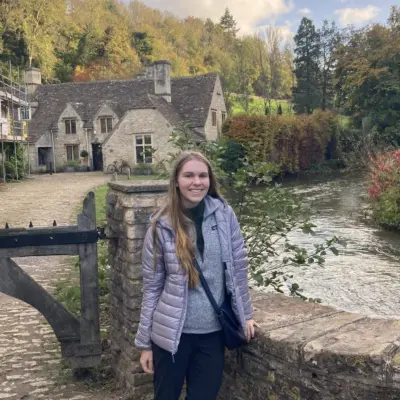 A woman with long blonde hair in a purple jacket stands in front of a cottage house along a river path.