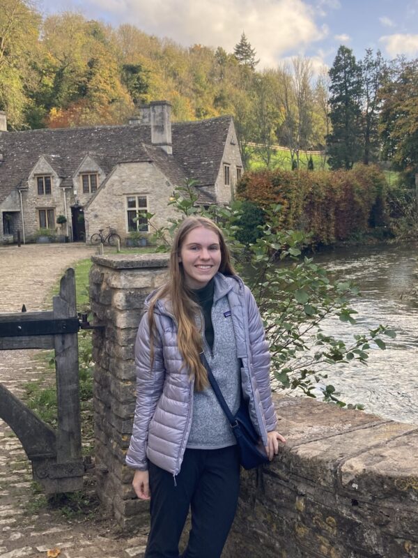 A woman with long blonde hair in a purple jacket stands in front of a cottage house along a river path.