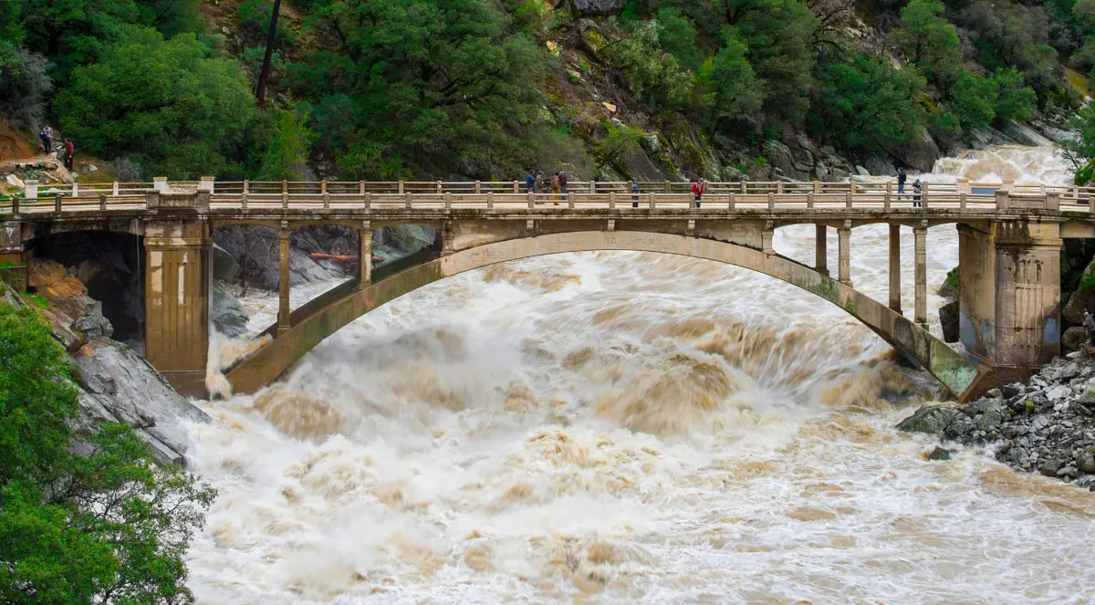 Muddy floodwaters surge through the arch of a concrete bridge in a forested river canyon as onlookers watch from above.