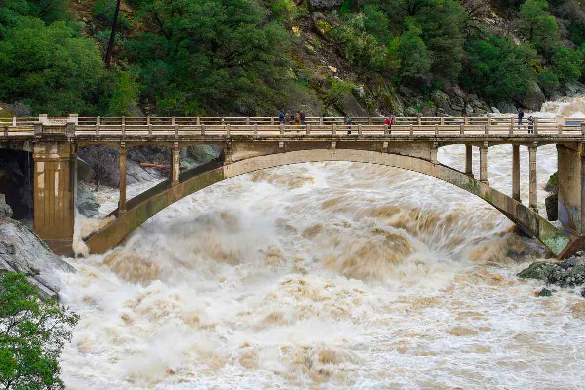 Muddy floodwaters surge through the arch of a concrete bridge in a forested river canyon as onlookers watch from above.