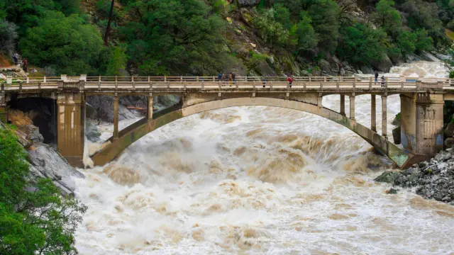 Muddy floodwaters surge through the arch of a concrete bridge in a forested river canyon as onlookers watch from above.