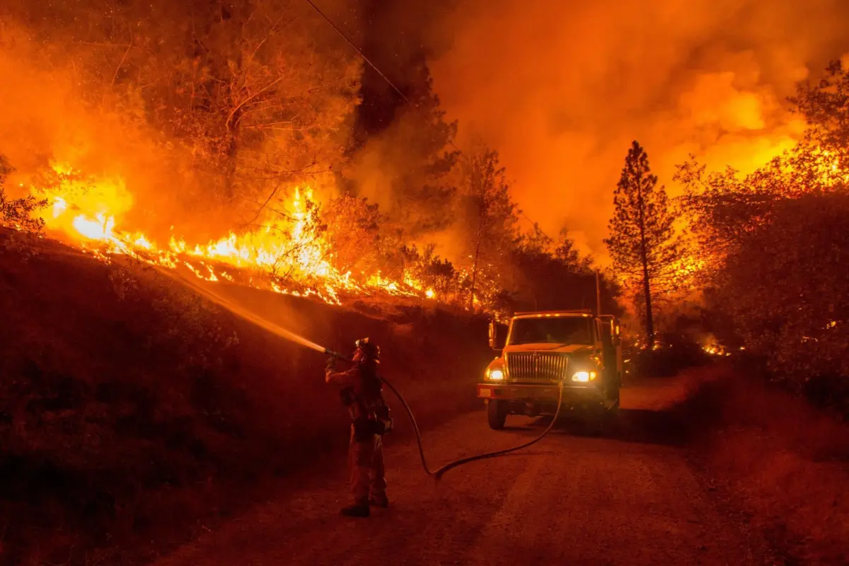 A firefighter aims a hose at flames burning up a hillside beside a dirt road at night, with a fire engine nearby.