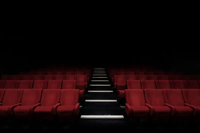 Empty rows of red theater seats flanking a lit center aisle stairway in a darkened auditorium.