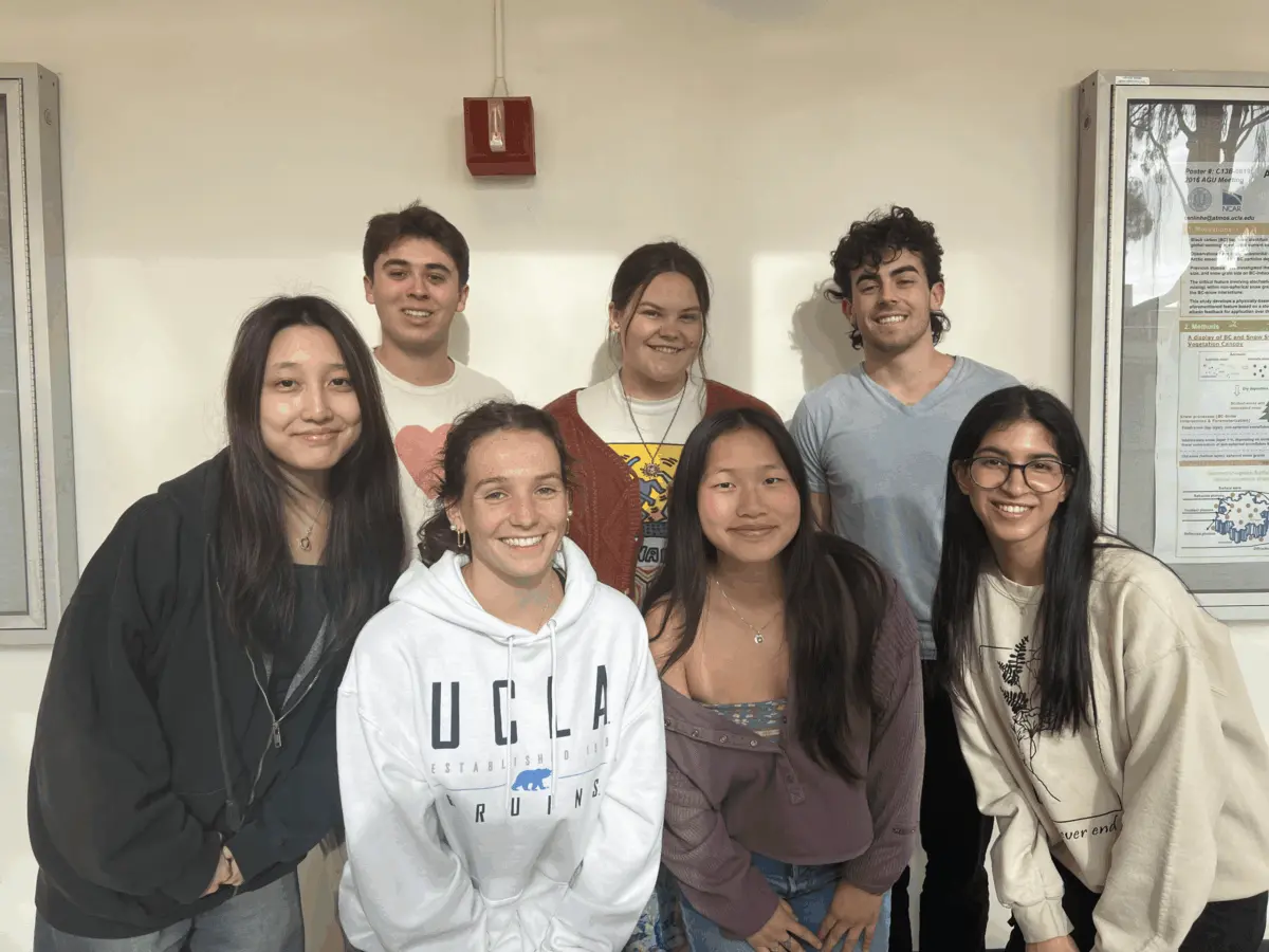 A group of seven UCLA students gathered together indoors beside a bulletin board.