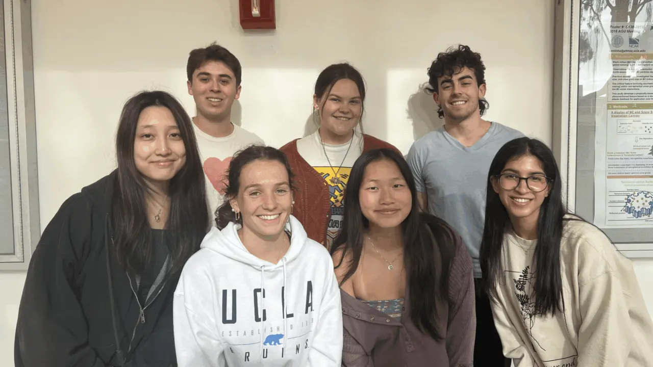 A group of seven UCLA students gathered together indoors beside a bulletin board.
