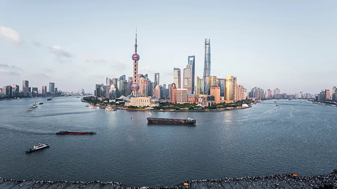 Shanghai's Pudong skyline across the Huangpu River, with cargo barges and boats passing and crowds along the waterfront promenade.