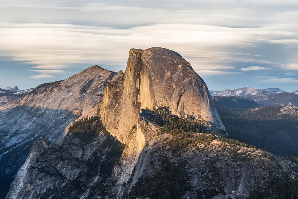Half Dome rises above forested ridges in Yosemite National Park, its granite face lit by low sun under streaked clouds.