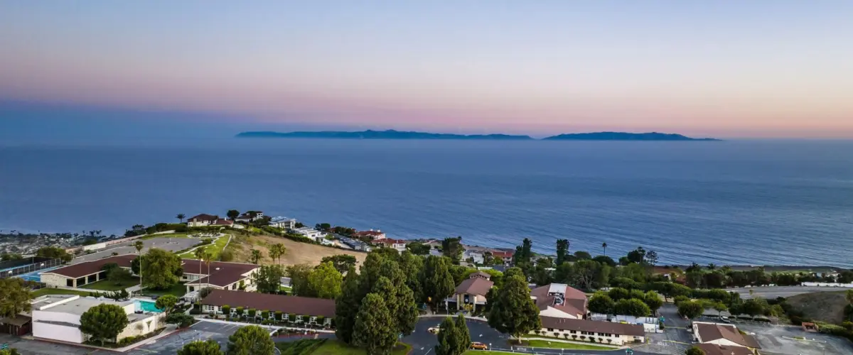 Aerial view of a coastal campus and parking lots above the ocean at dusk, with an island visible on the horizon.