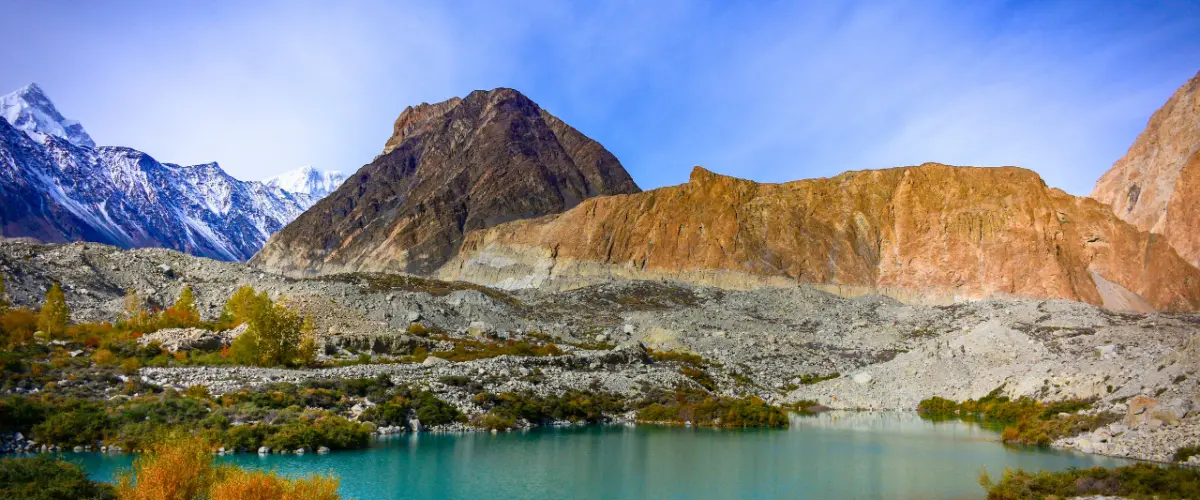 A turquoise glacial lake ringed by autumn shrubs below rocky peaks and snowcapped mountains under a deep blue sky.