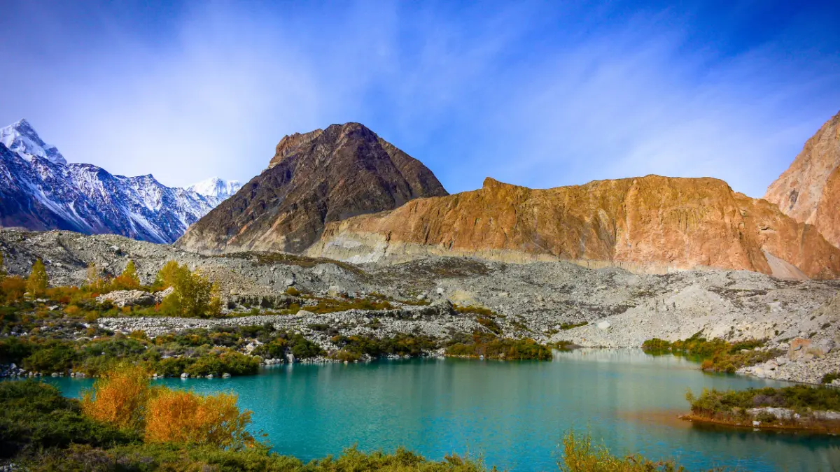 A turquoise glacial lake ringed by autumn shrubs below rocky peaks and snowcapped mountains under a deep blue sky.
