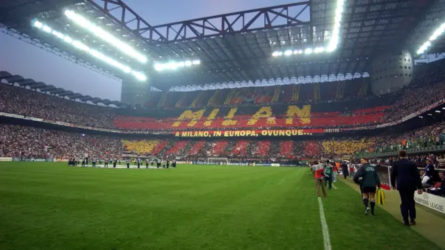Packed stadium at San Siro where fans in the stands form a red and black display spelling "Milan" above the pitch before a match.