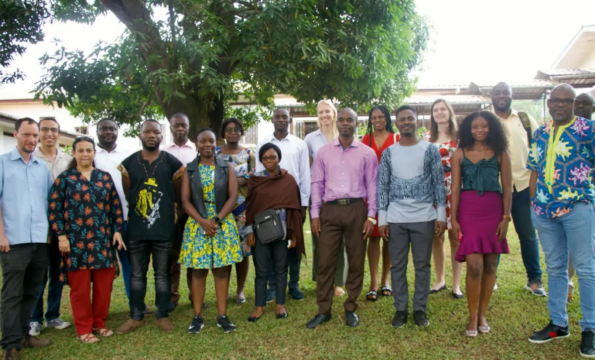 A large group of workshop participants stands together for a group portrait on a lawn beneath a shade tree.