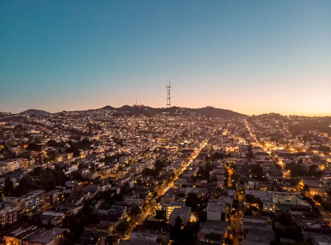 Aerial view of San Francisco neighborhoods at dusk, with street lights glowing and Sutro Tower on the hilltop against a clear sky.