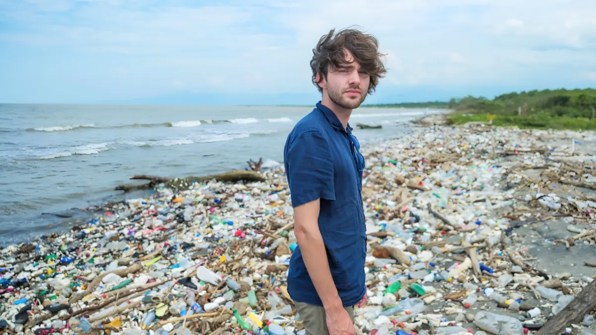A person stands on a shoreline blanketed in plastic bottles and debris washed up along the water's edge.