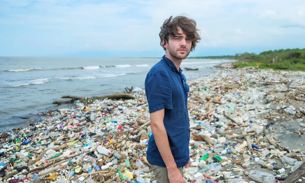 A person stands on a shoreline blanketed in plastic bottles and debris washed up along the water's edge.