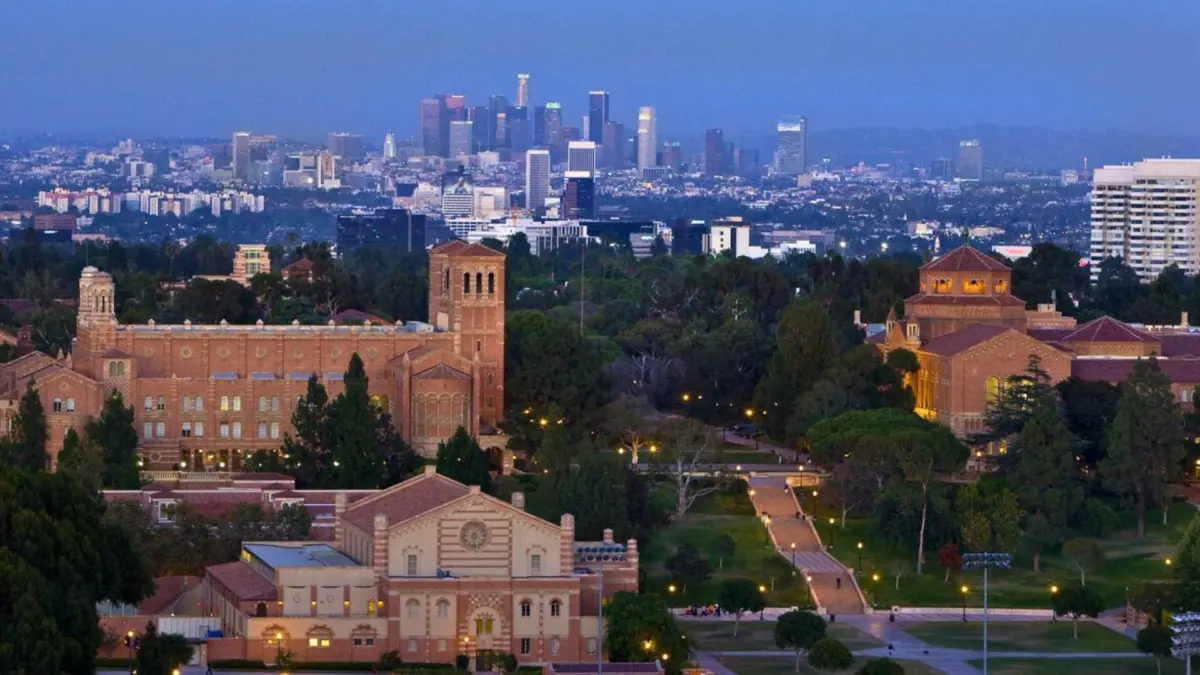UCLA's campus buildings lit at dusk, with the downtown Los Angeles skyline rising in the distance.