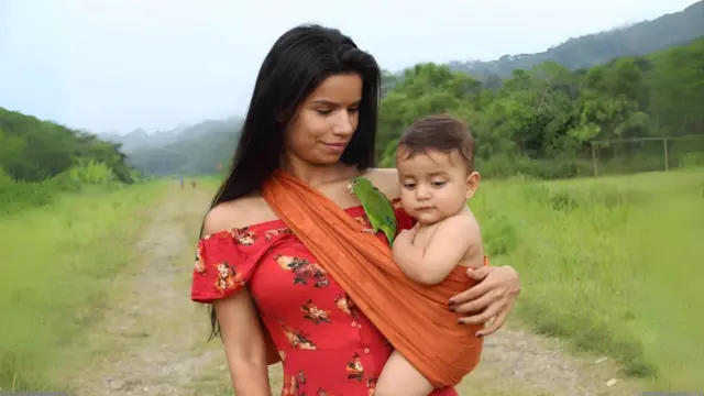 A woman carries a baby in a sling along a grassy dirt path in a forested valley, with a small green parrot perched on the sling.