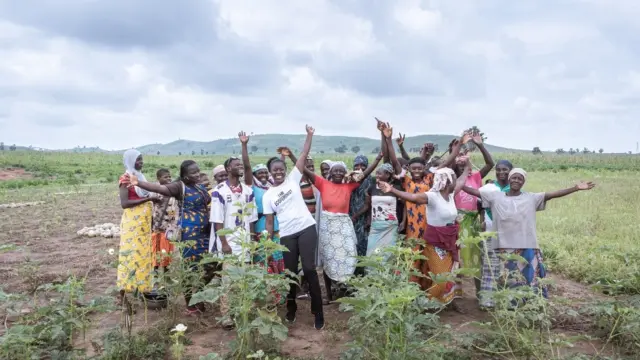 A group of women stand together in an open field, raising their arms and cheering under a cloudy sky.