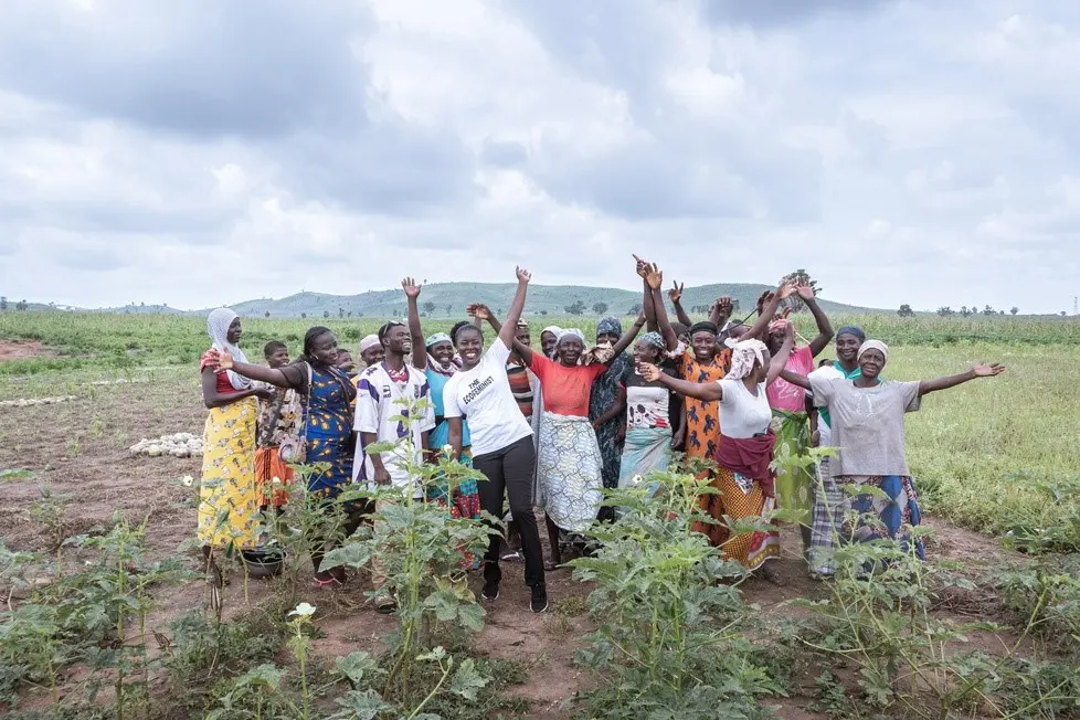 A group of women stand together in an open field, raising their arms and cheering under a cloudy sky.