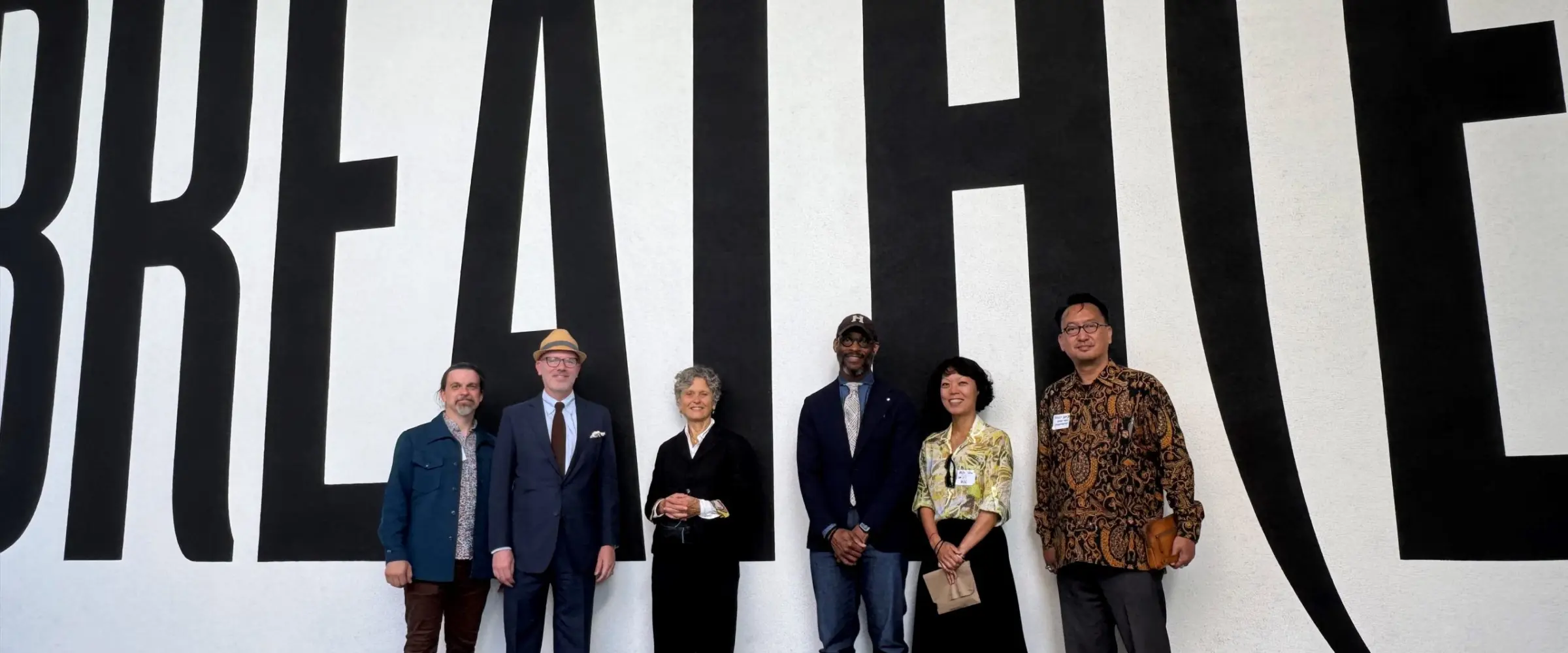 Six workshop participants stand together in front of a large wall mural spelling out "BREATH(E)" in oversized black letters.