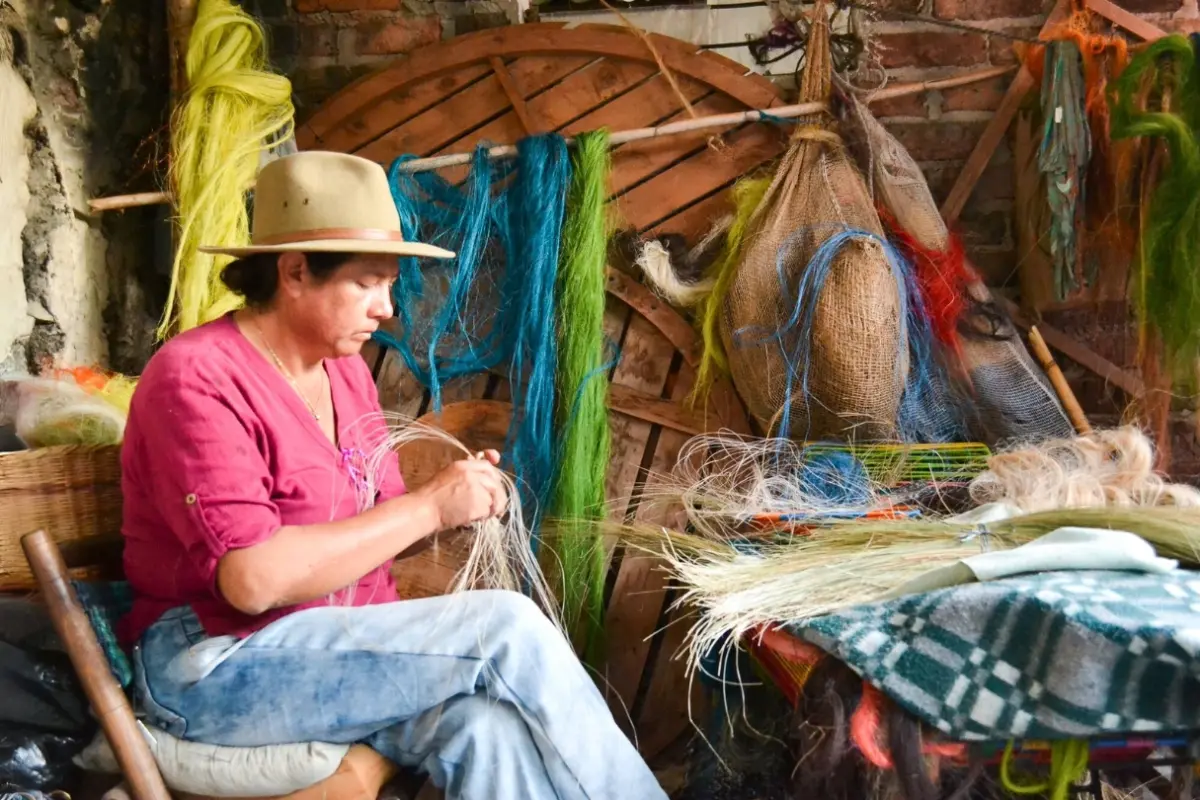 Angelina Sanchez Bustacara hand weaves plant fibers in a workshop surrounded by dyed fiber bundles and baskets.