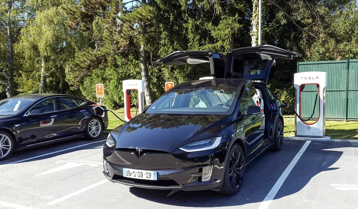 A Tesla Model X with its falcon-wing doors raised charges at a Tesla Supercharger station, with another Tesla parked nearby.