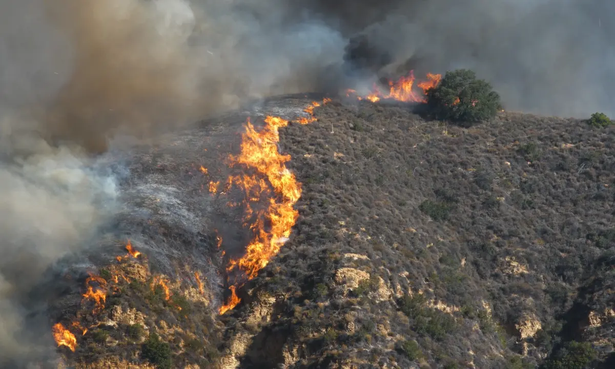Flames spread up a brush-covered hillside as thick smoke billows over the ridge during a wildfire.