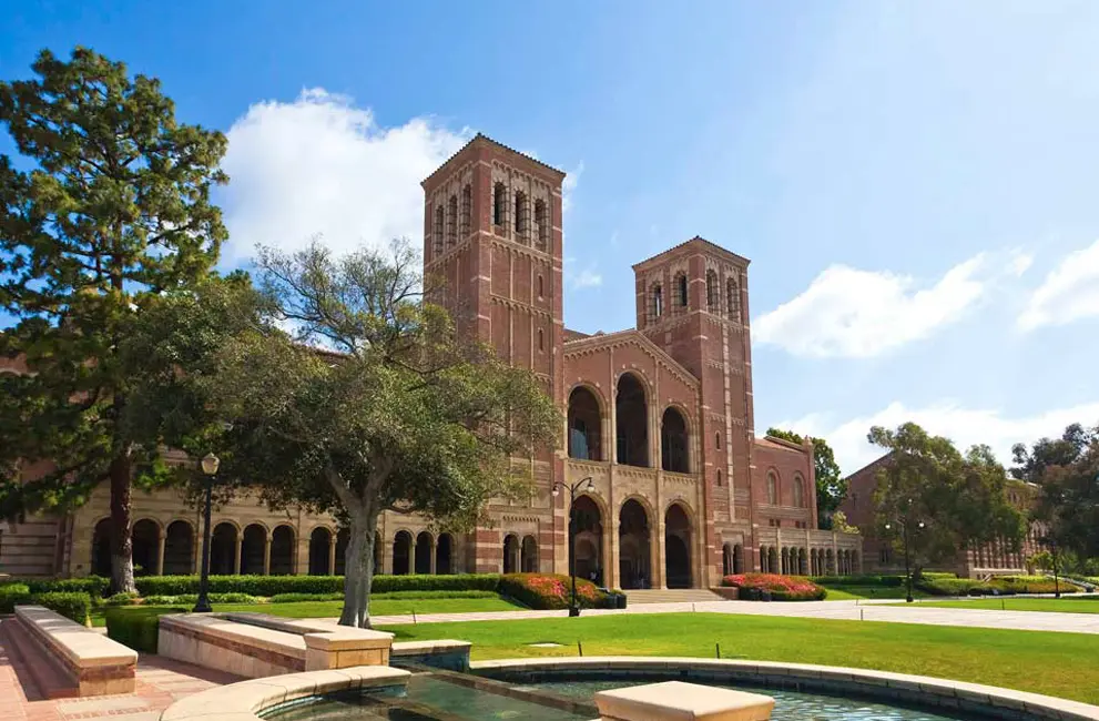 UCLA's Royce Hall with its twin brick towers, seen across a lawn and fountain under a blue sky.