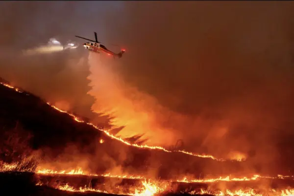 A helicopter drops water on a hillside wildfire at night, with flames spreading along the ridge.