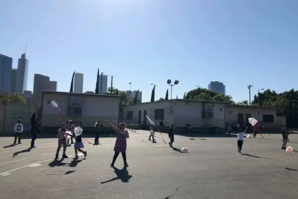Children fly kites on a paved schoolyard beside portable classrooms, with the downtown Los Angeles skyline in the background.