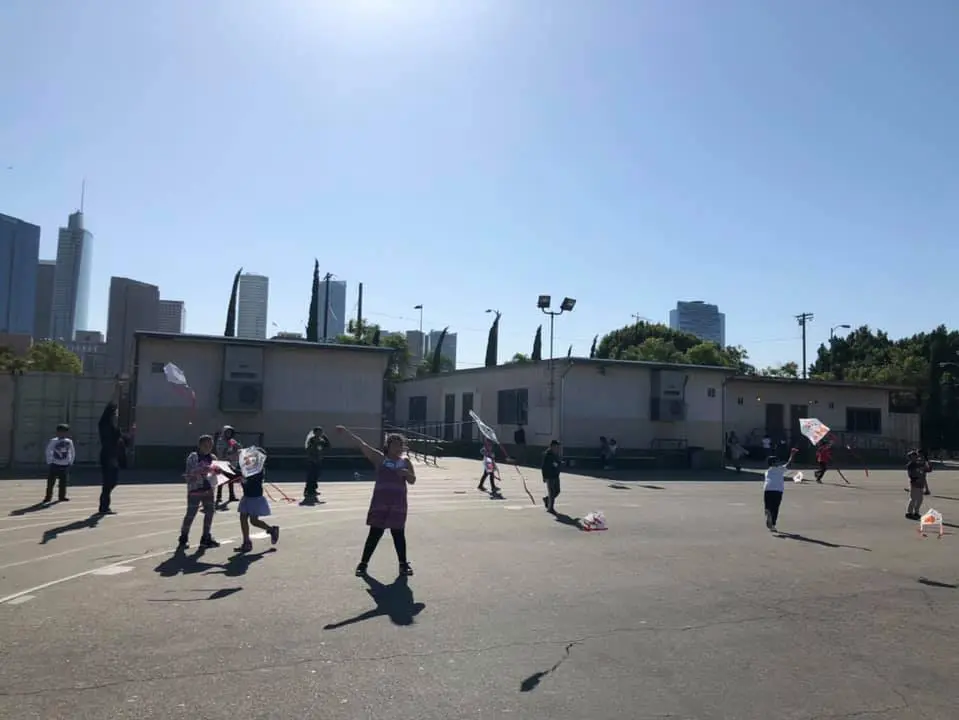 Children fly kites on a paved schoolyard beside portable classrooms, with the downtown Los Angeles skyline in the background.