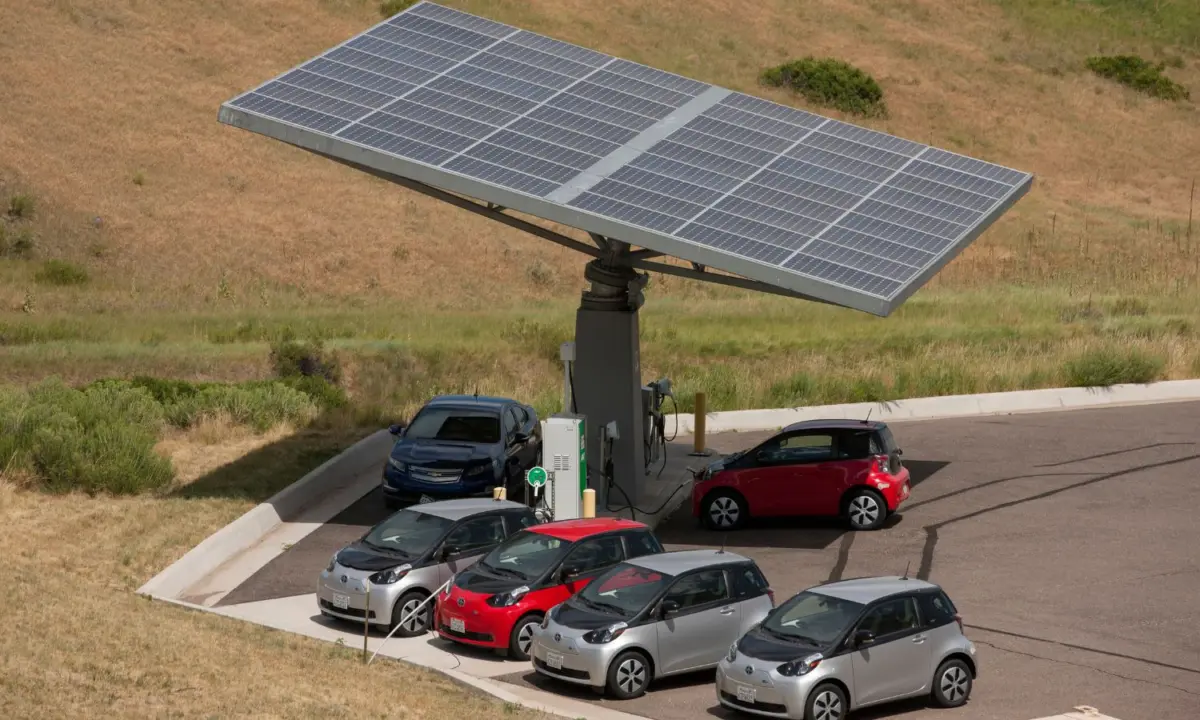 Electric cars parked at a charging station shaded by a large solar panel array on a dry grassy hillside.