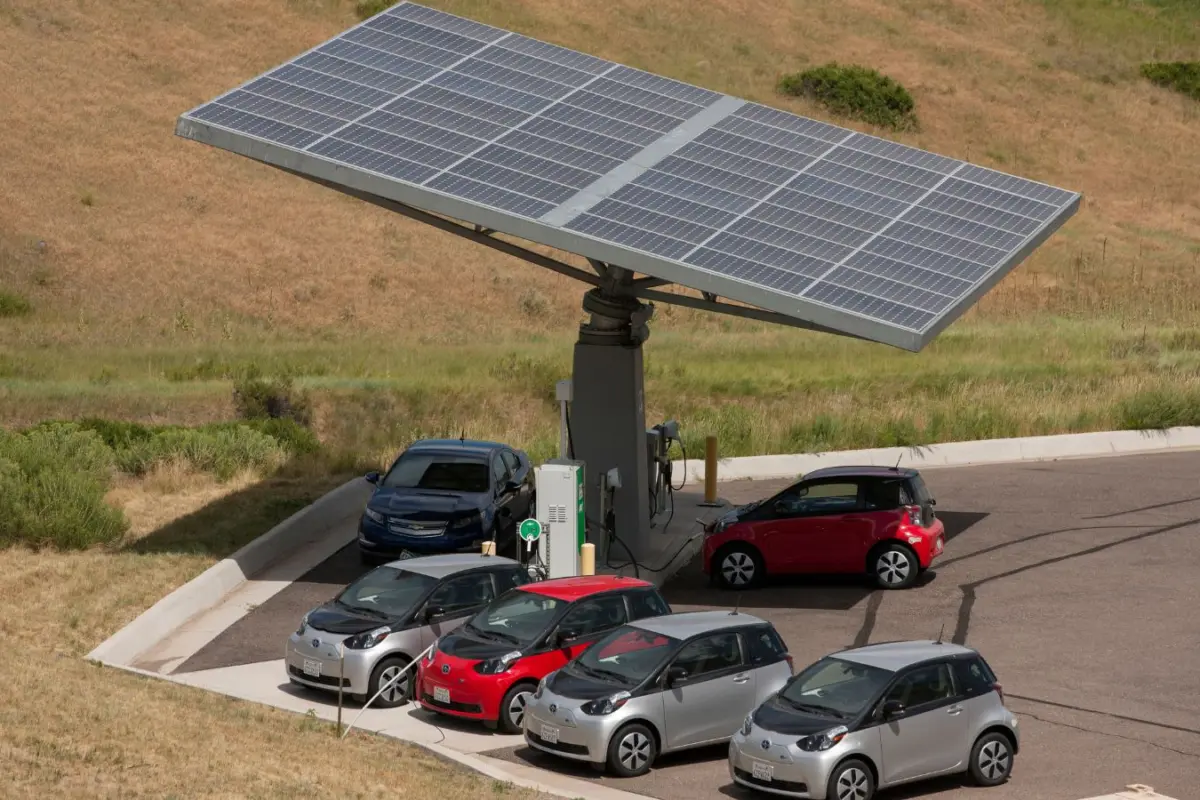 Electric cars parked at a charging station shaded by a large solar panel array on a dry grassy hillside.