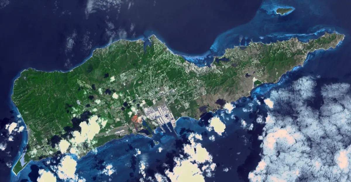 Satellite view of St. Croix, a green island ringed by shallow reefs and deep blue ocean, with scattered clouds and a developed coastal area.