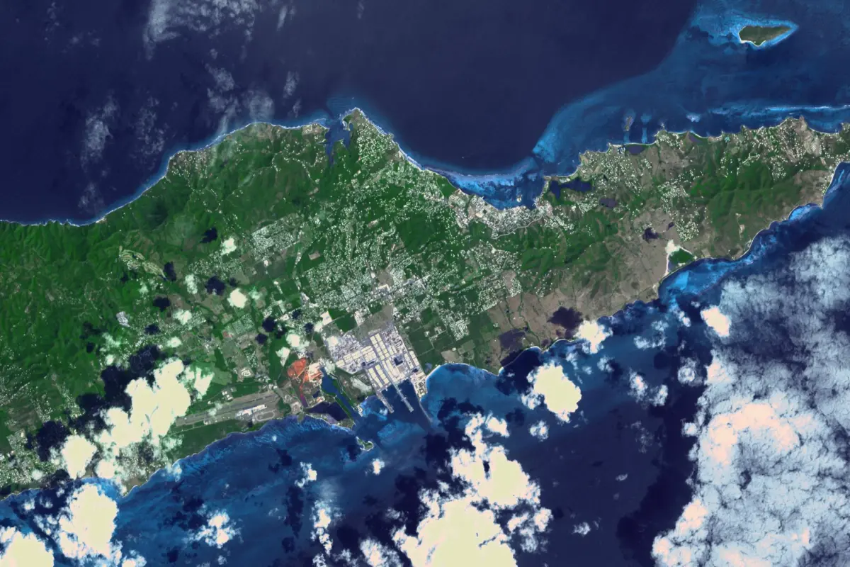 Satellite view of St. Croix, a green island ringed by shallow reefs and deep blue ocean, with scattered clouds and a developed coastal area.