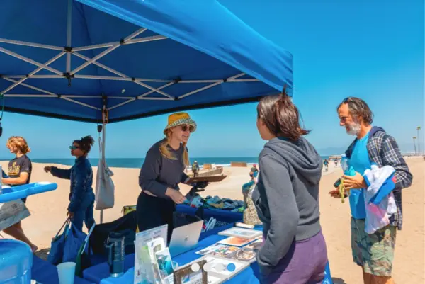 Volunteers talk with beach visitors at an outreach tent set up on the sand during a beach cleanup event.