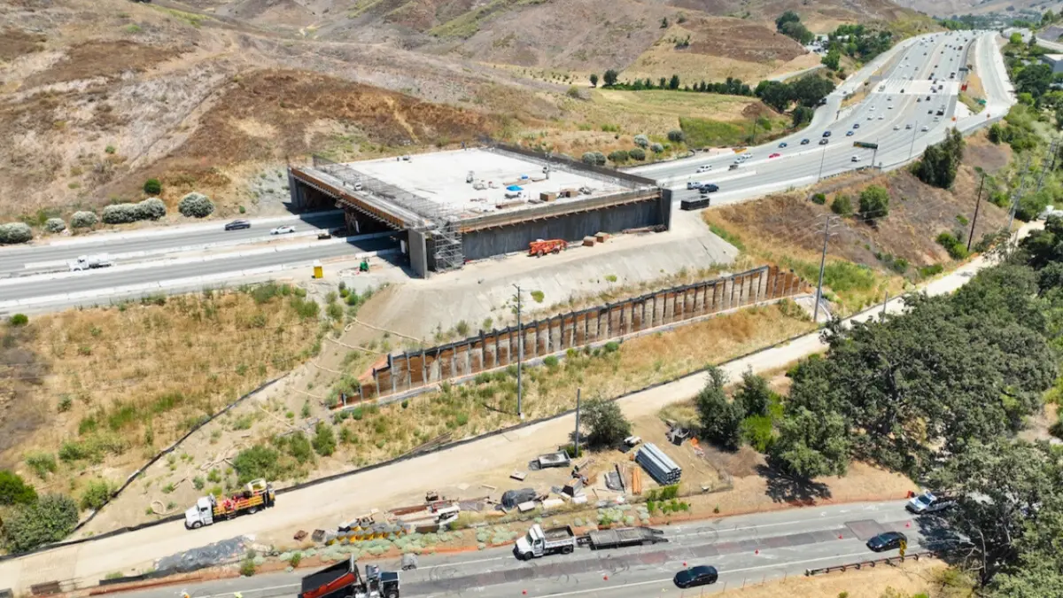 Aerial view of the Wallis Annenberg Wildlife Crossing under construction, spanning 10 lanes of freeway amid hills and oak trees.
