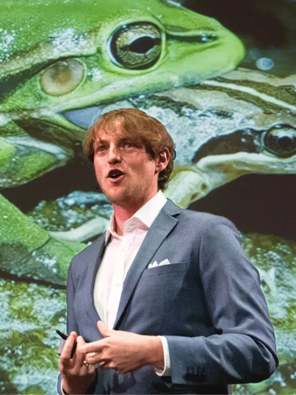 Anthony Waddle speaking on stage in front of a large projected image of frogs
