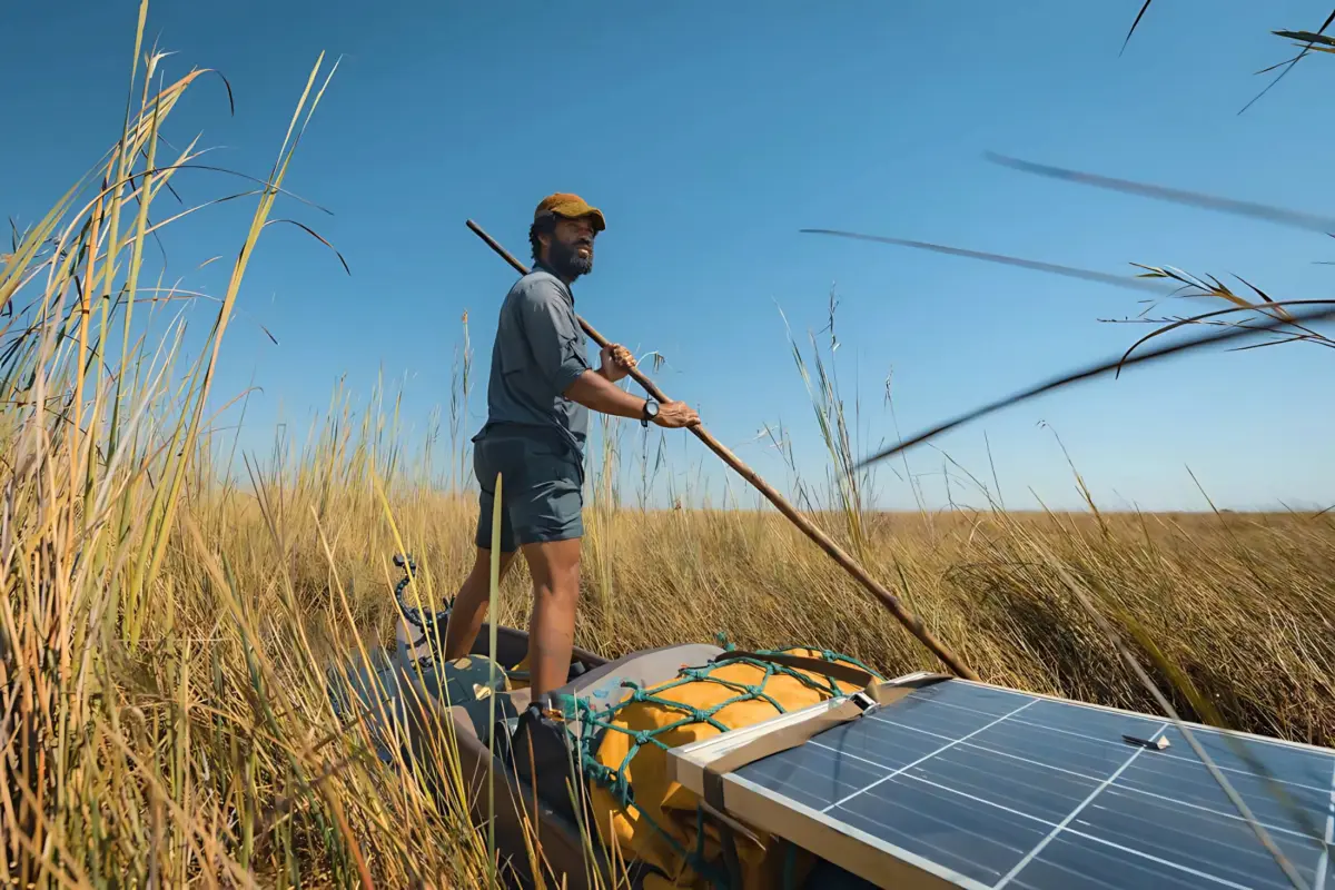 Kerllen da Silva Freitas Costa stands on a narrow canoe outfitted with solar panels, steering through tall grasses in a remote wetland under a clear blue sky.