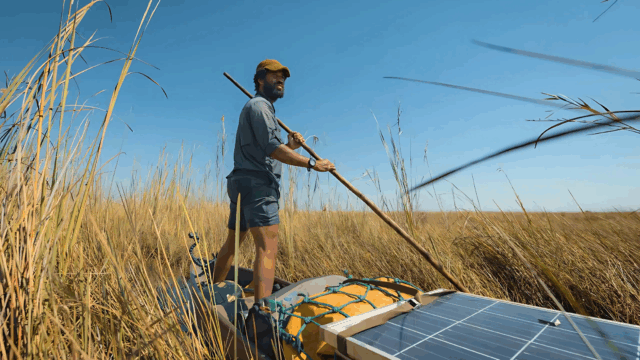 Kerllen da Silva Freitas Costa stands on a narrow canoe outfitted with solar panels, steering through tall grasses in a remote wetland under a clear blue sky.