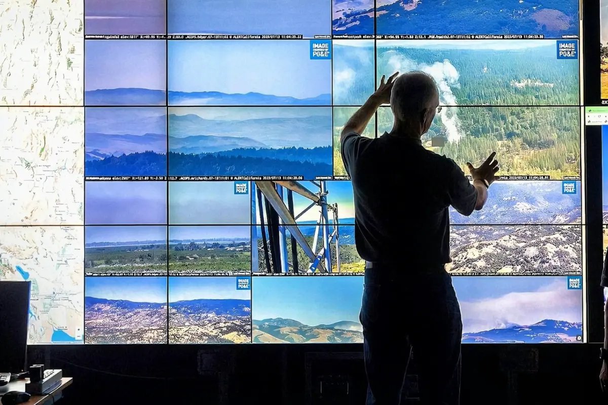 Two people stand in front of a large multi-screen display wall showing real-time wildfire camera feeds and a map of California. One person gestures toward the screens, analyzing smoke and fire activity.