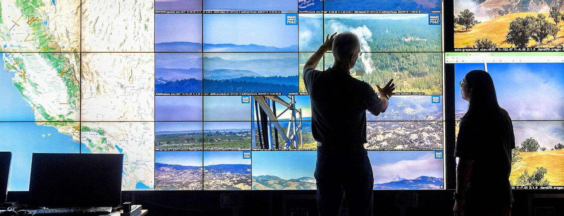Two people stand in front of a large multi-screen display wall showing real-time wildfire camera feeds and a map of California. One person gestures toward the screens, analyzing smoke and fire activity.