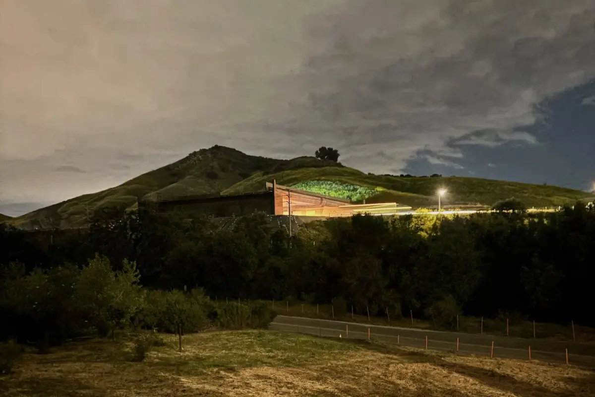 Image of a road at night with a bridge going over the road and lights emanating from under the bridge. More light is seen across a field in the foreground from another source. There is a hill in the background and the clouds above are reflecting light as well.