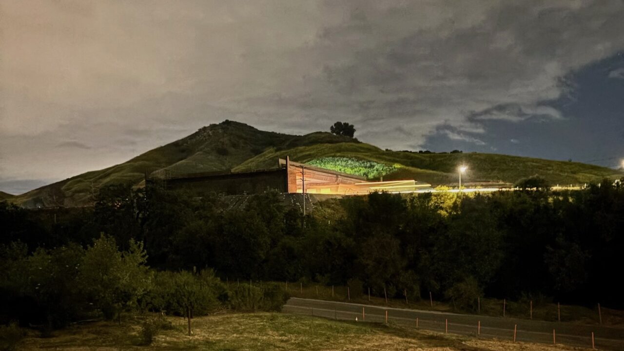 Image of a road at night with a bridge going over the road and lights emanating from under the bridge. More light is seen across a field in the foreground from another source. There is a hill in the background and the clouds above are reflecting light as well.