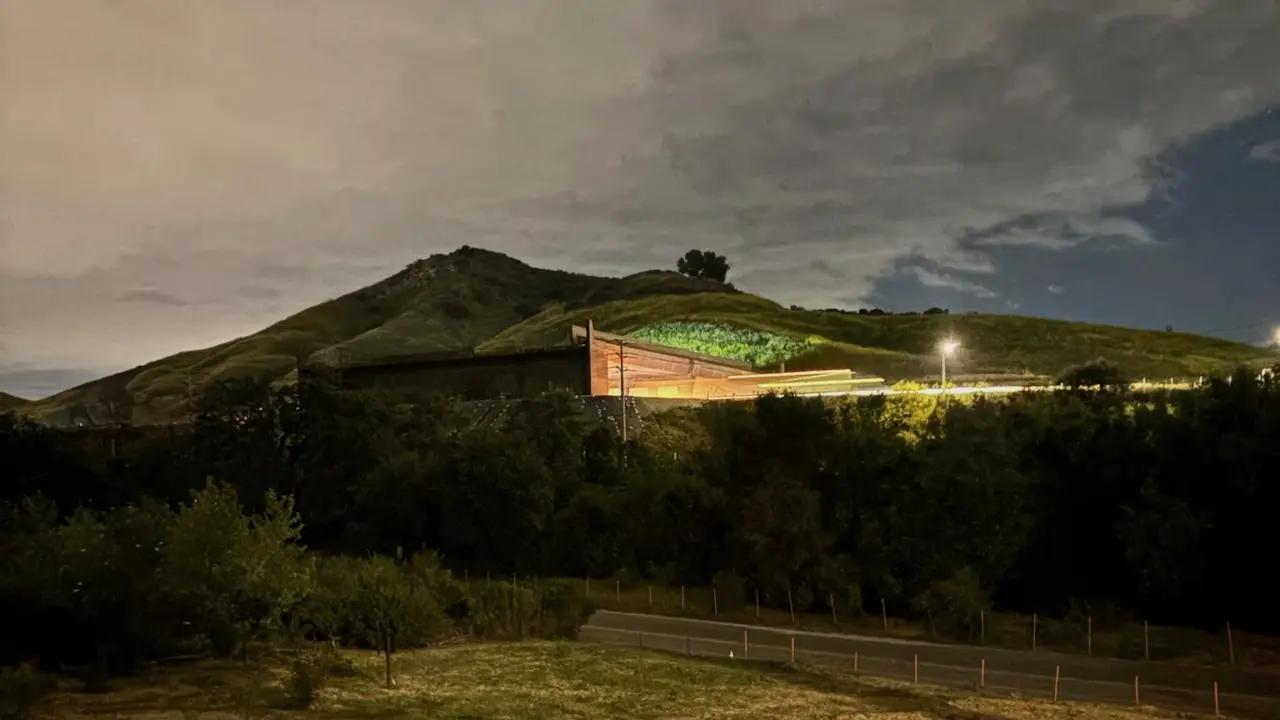 Image of a road at night with a bridge going over the road and lights emanating from under the bridge. More light is seen across a field in the foreground from another source. There is a hill in the background and the clouds above are reflecting light as well.