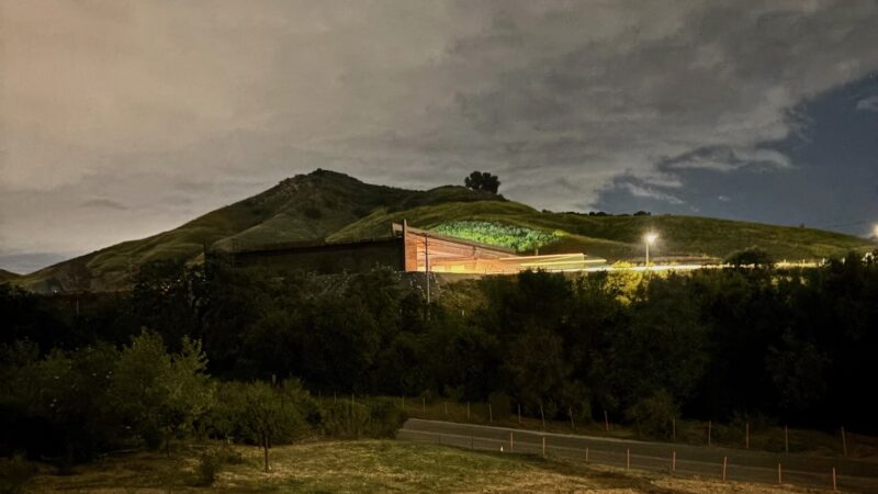 Image of a road at night with a bridge going over the road and lights emanating from under the bridge. More light is seen across a field in the foreground from another source. There is a hill in the background and the clouds above are reflecting light as well.