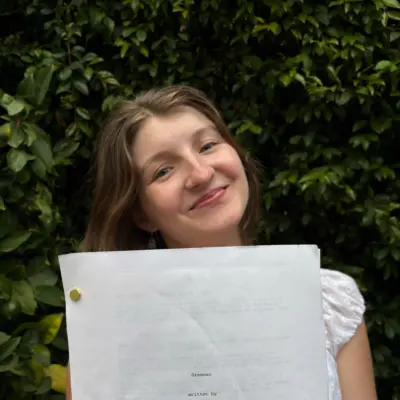 Emily McFerson holding up a printed script she wrote, in front of a leafy hedge.