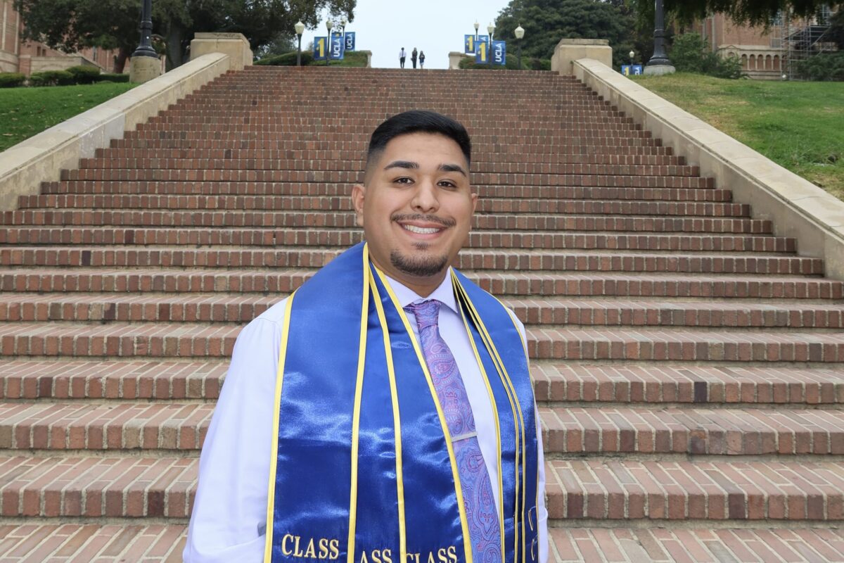 Marcos Magana, a Latino man with short black hair, stands smiling in front of UCLA’s Janss Steps, wearing a white button-up shirt, black tie, and blue graduation sash.