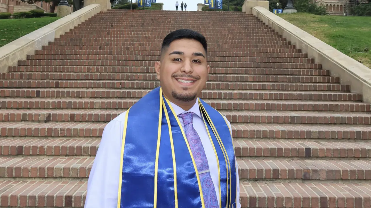 Marcos Magana, a Latino man with short black hair, stands smiling in front of UCLA’s Janss Steps, wearing a white button-up shirt, black tie, and blue graduation sash.
